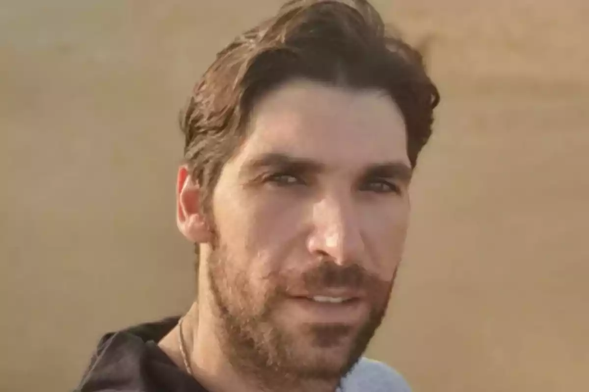 Un hombre con barba y cabello oscuro mira a la cámara con una expresión neutral y un fondo desenfocado.