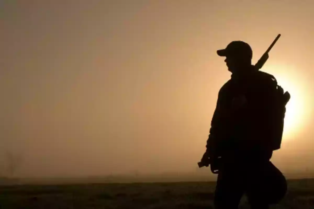 Silueta de una persona con gorra y rifle al hombro al amanecer.