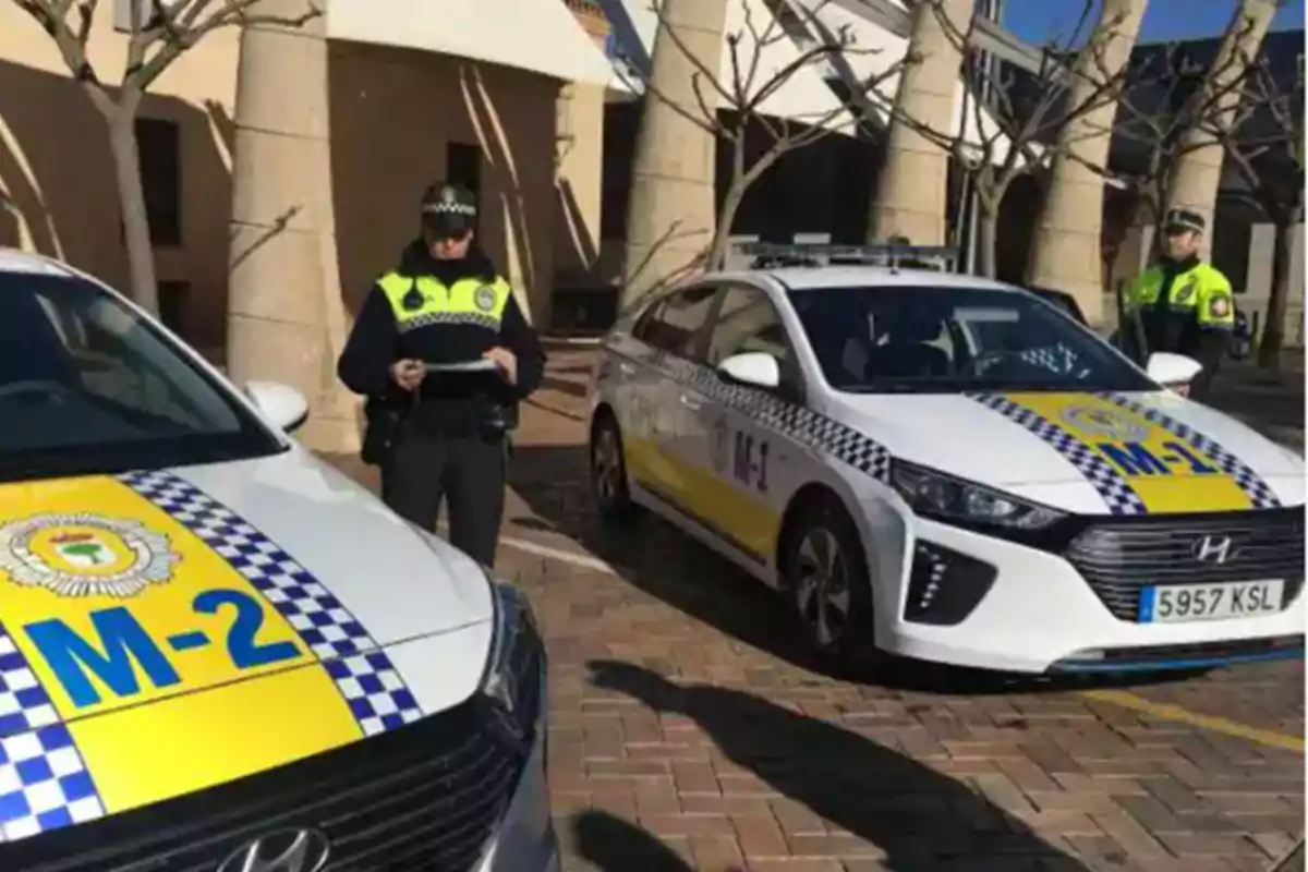 Dos agentes de policía están de pie junto a dos coches patrulla con distintivos amarillos y azules en una plaza pavimentada.