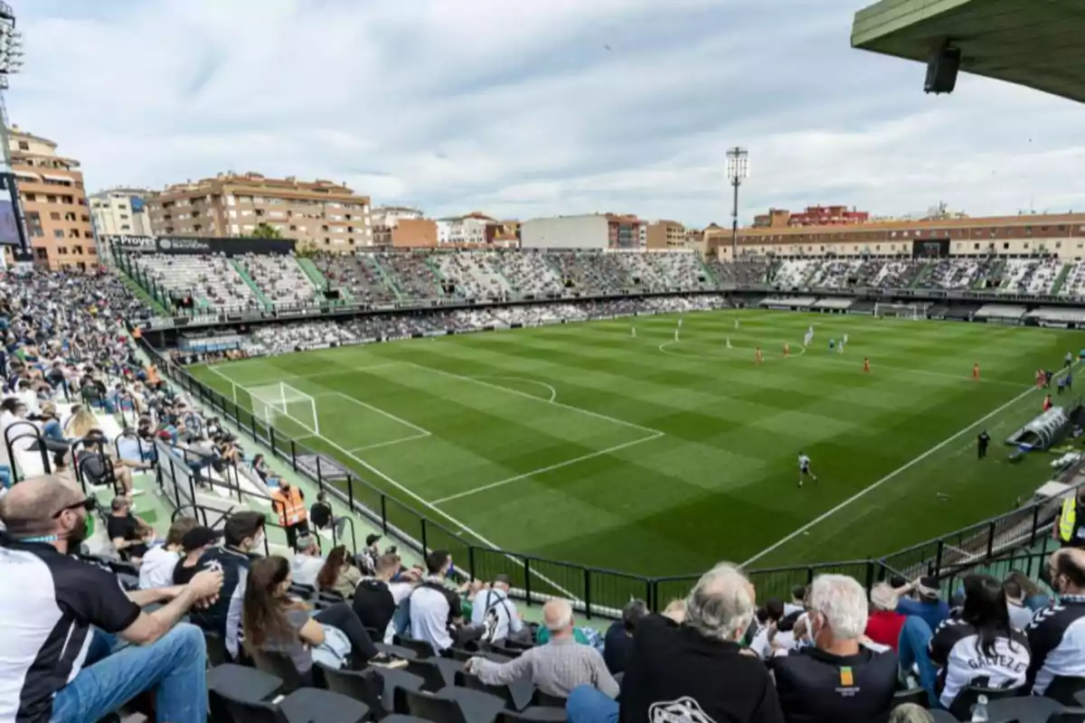 Un estadio de fútbol lleno de espectadores con un partido en curso y edificios residenciales al fondo.