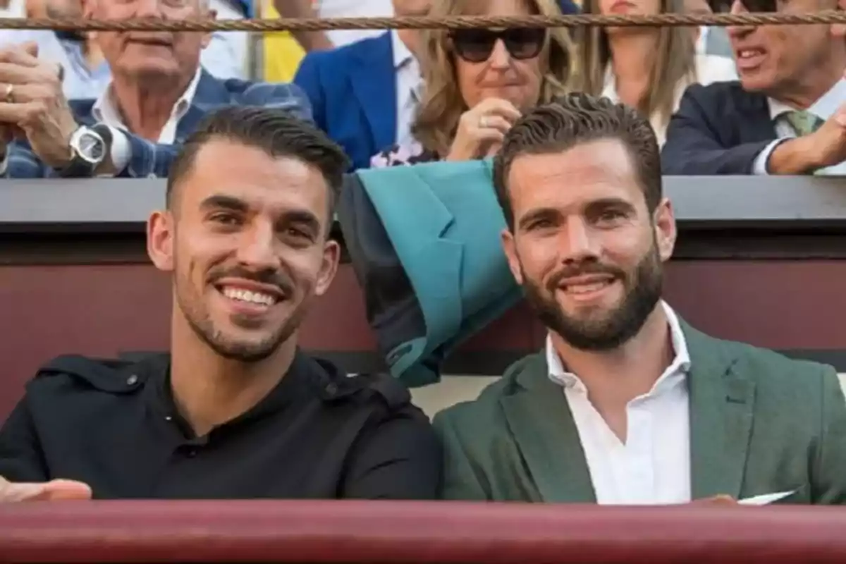 Dos hombres sonrientes sentados en un evento, con personas en el fondo.