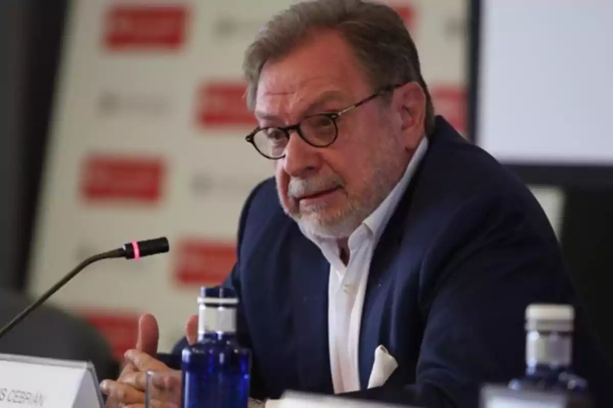 Un hombre con gafas y barba hablando en un micrófono durante un evento con una botella de agua en la mesa.