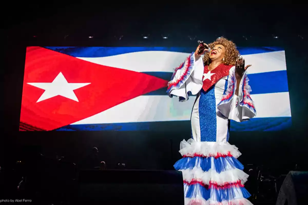 Una cantante vestida con un traje colorido y patriótico canta en un escenario frente a una gran pantalla que muestra la bandera de Cuba.