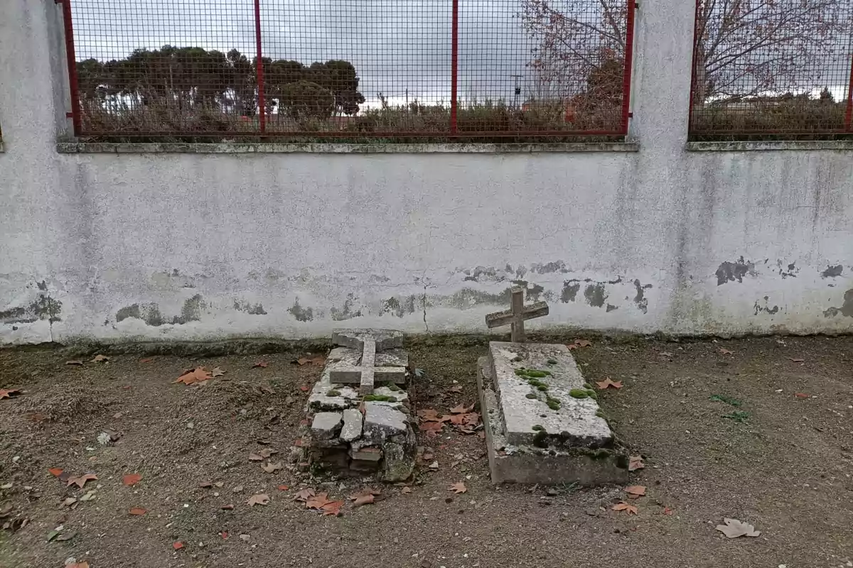 Dos tumbas antiguas con cruces de piedra se encuentran junto a una pared desgastada y un enrejado rojo, rodeadas de hojas secas y musgo, bajo un cielo nublado. Dos tumbas antiguas con cruces de piedra se encuentran junto a una pared desgastada y un enrejado rojo, rodeadas de hojas secas y musgo, bajo un cielo nublado.