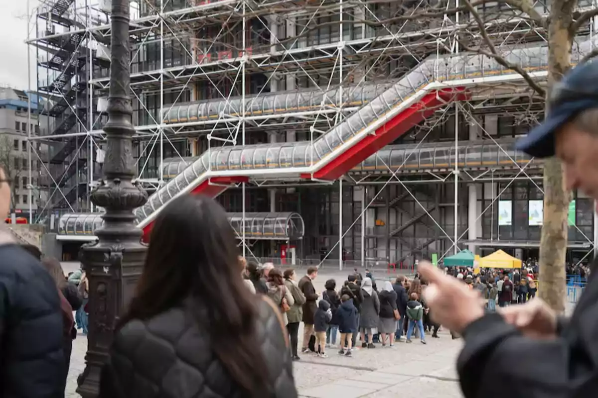 Personas haciendo fila frente a un edificio moderno con una estructura de tubos y escaleras mecánicas visibles en el exterior.