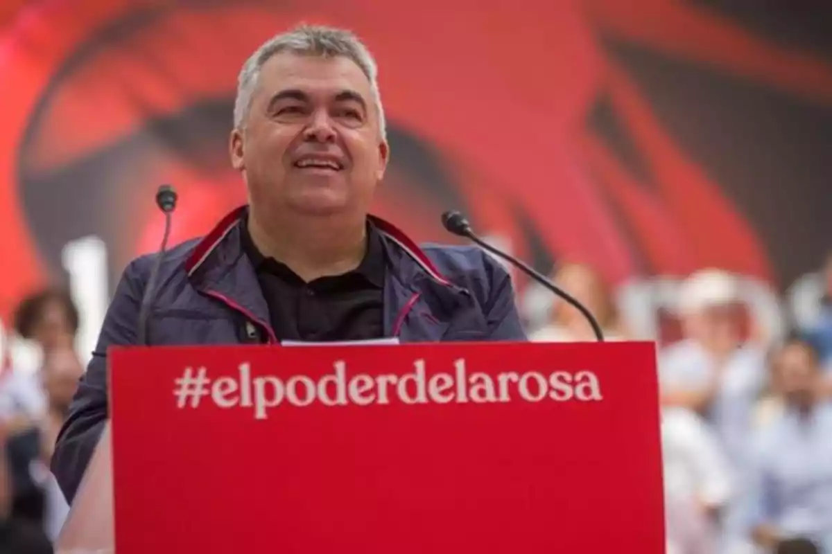 Un hombre sonriente habla en un podio rojo con el texto "#elpoderdelarosa" y un fondo desenfocado.