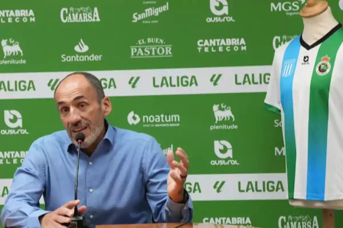 Un hombre hablando en una conferencia de prensa con un micrófono frente a un fondo verde con logotipos de patrocinadores y una camiseta de fútbol en un maniquí a su lado.