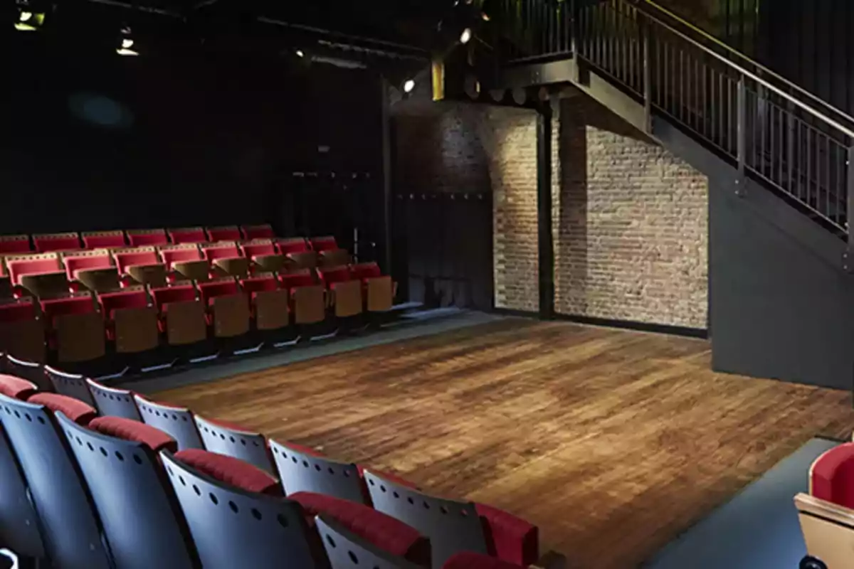 Un pequeño teatro con asientos rojos y negros frente a un escenario de madera, con una pared de ladrillo y una escalera metálica al fondo.