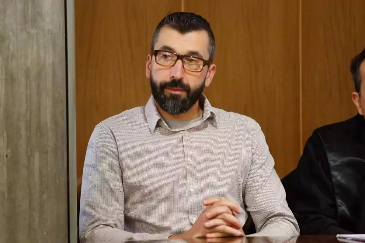 Un hombre con barba y gafas está sentado con las manos entrelazadas frente a una mesa en un entorno interior.