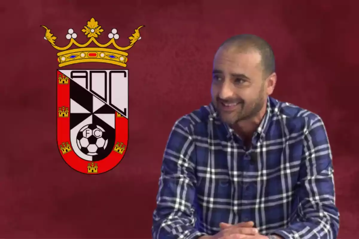 Un hombre con camisa a cuadros sonríe frente a un fondo rojo con un escudo heráldico a su lado.