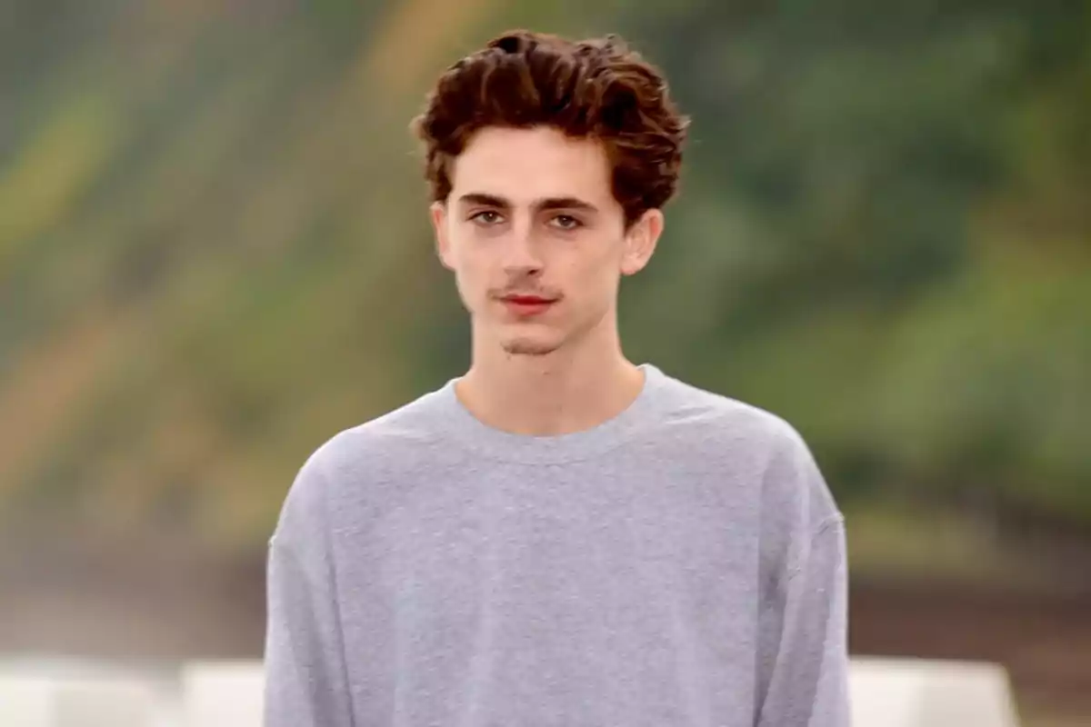 Un joven con cabello castaño y ondulado lleva una camiseta gris y está de pie frente a un fondo desenfocado.