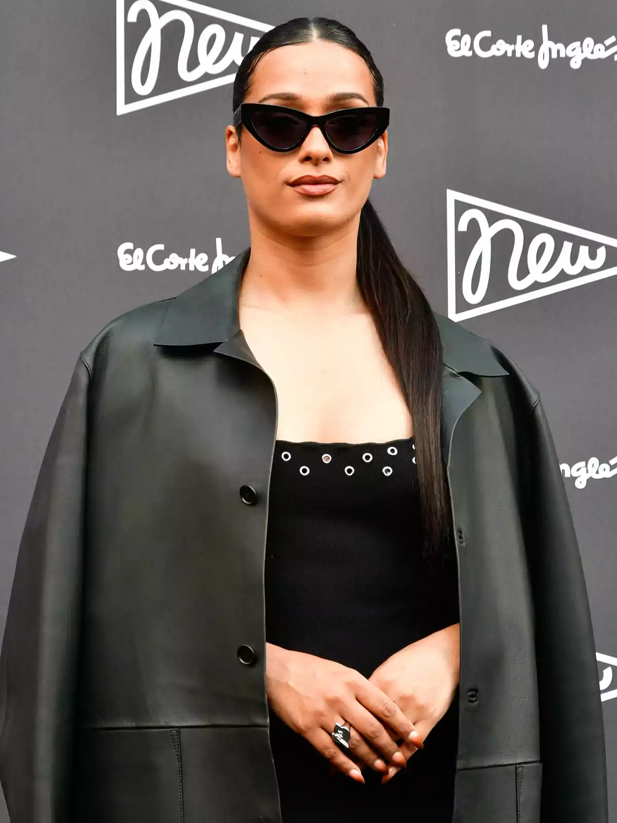 Chanel con gafas de sol y cabello largo recogido lleva un abrigo negro sobre un vestido negro con detalles metálicos, posando frente a un fondo con logotipos de El Corte Inglés. Chanel con gafas de sol y cabello largo recogido lleva un abrigo negro sobre un vestido negro con detalles metálicos, posando frente a un fondo con logotipos de El Corte Inglés.