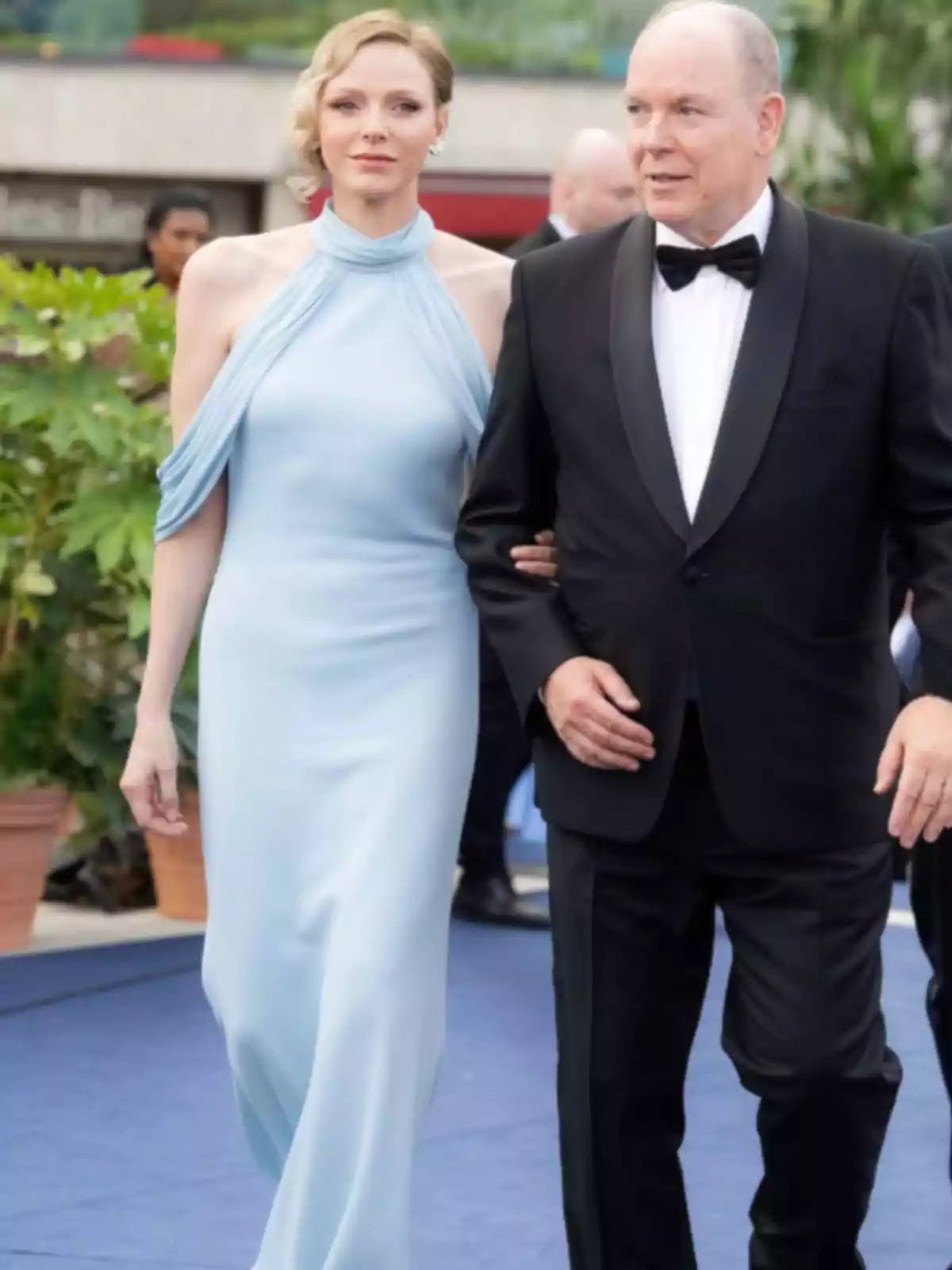 Una mujer con vestido azul claro y un hombre con esmoquin negro caminan juntos por una alfombra azul en un evento elegante al aire libre. Una mujer con vestido azul claro y un hombre con esmoquin negro caminan juntos por una alfombra azul en un evento elegante al aire libre.