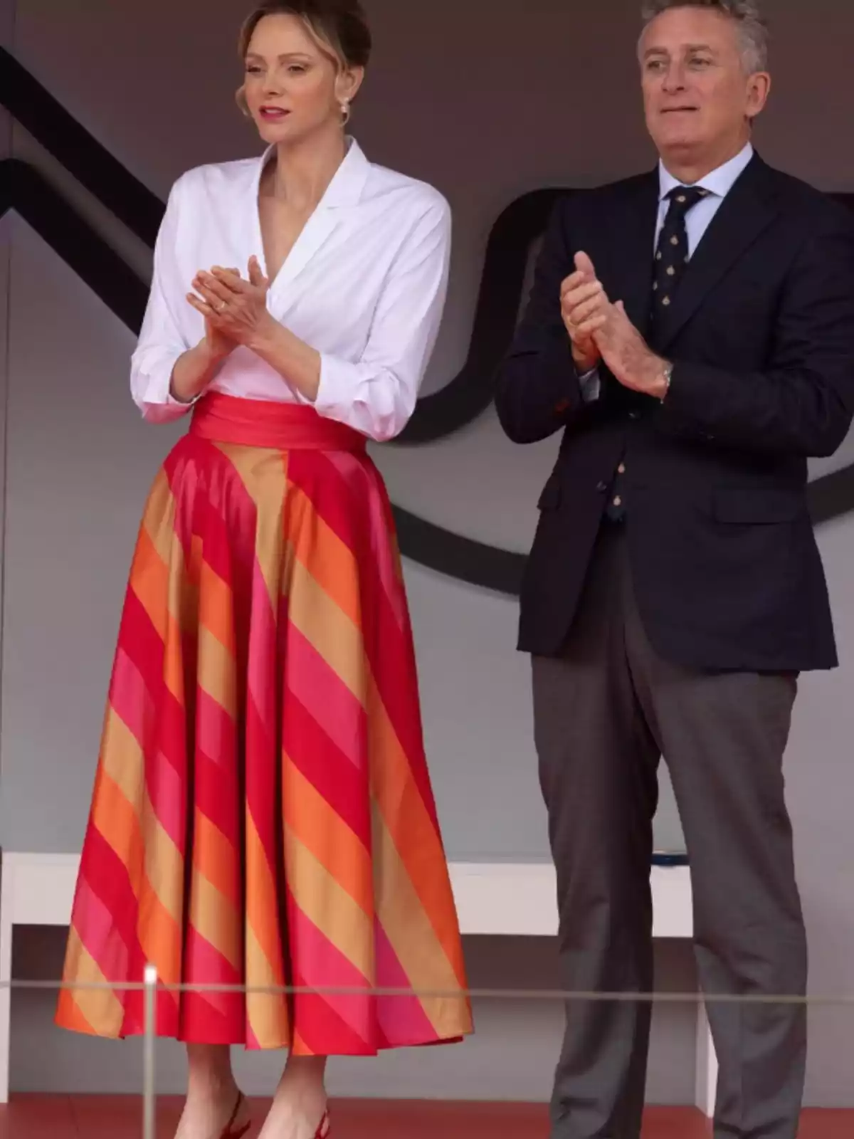 Una mujer con blusa blanca y falda de rayas rojas y naranjas está de pie junto a un hombre con traje oscuro, ambos aplaudiendo. Una mujer con blusa blanca y falda de rayas rojas y naranjas está de pie junto a un hombre con traje oscuro, ambos aplaudiendo.