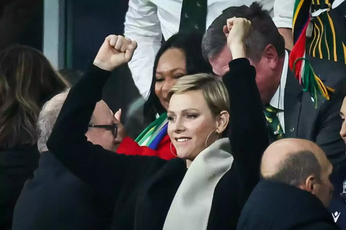 Charlene sonriendo con los brazos arriba, jersey negro y bufanda gris Charlene sonriendo con los brazos arriba, jersey negro y bufanda gris