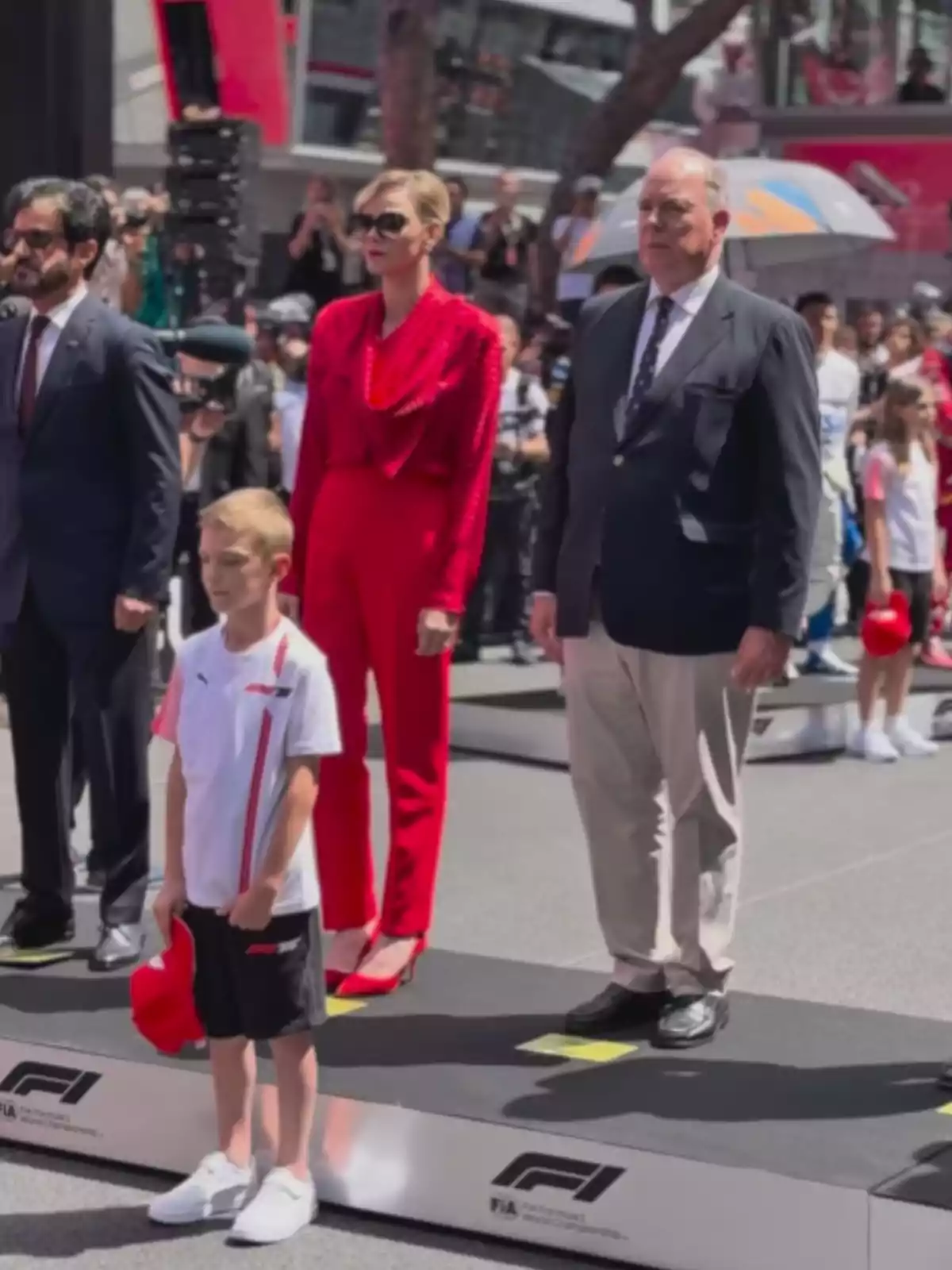Un grupo de personas está de pie en una plataforma durante un evento de Fórmula 1, con un niño al frente sosteniendo una gorra roja y una mujer vestida de rojo junto a un hombre con traje. Un grupo de personas está de pie en una plataforma durante un evento de Fórmula 1, con un niño al frente sosteniendo una gorra roja y una mujer vestida de rojo junto a un hombre con traje.