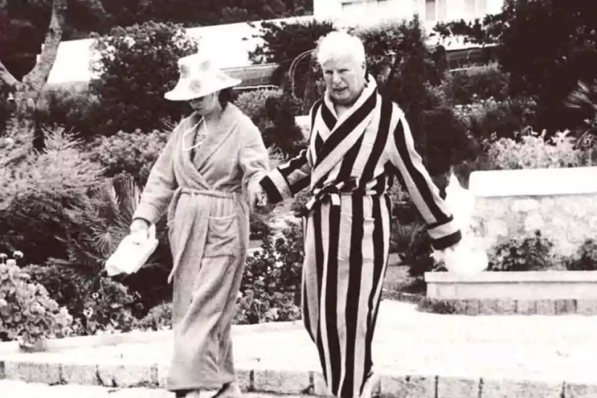 Una pareja de ancianos camina de la mano en un jardín, ambos vestidos con batas de baño, uno de ellos con una bata de rayas y el otro con una bata lisa y un sombrero.