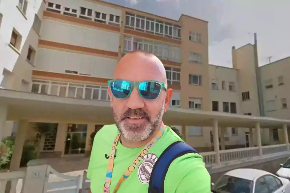 Hombre con gafas de sol y camiseta verde posando frente a un edificio de apartamentos.