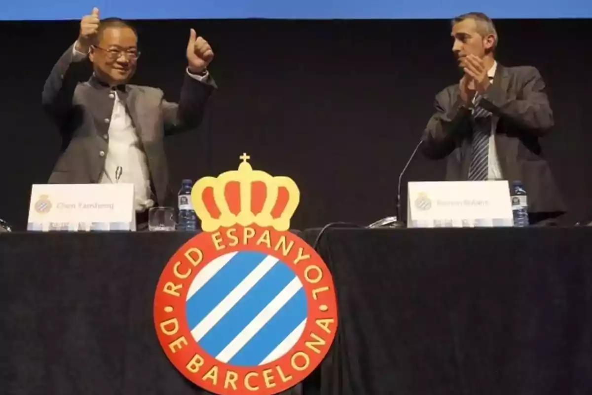 Dos hombres en un escenario con el logo del RCD Espanyol de Barcelona, uno de ellos levanta los pulgares mientras el otro aplaude.