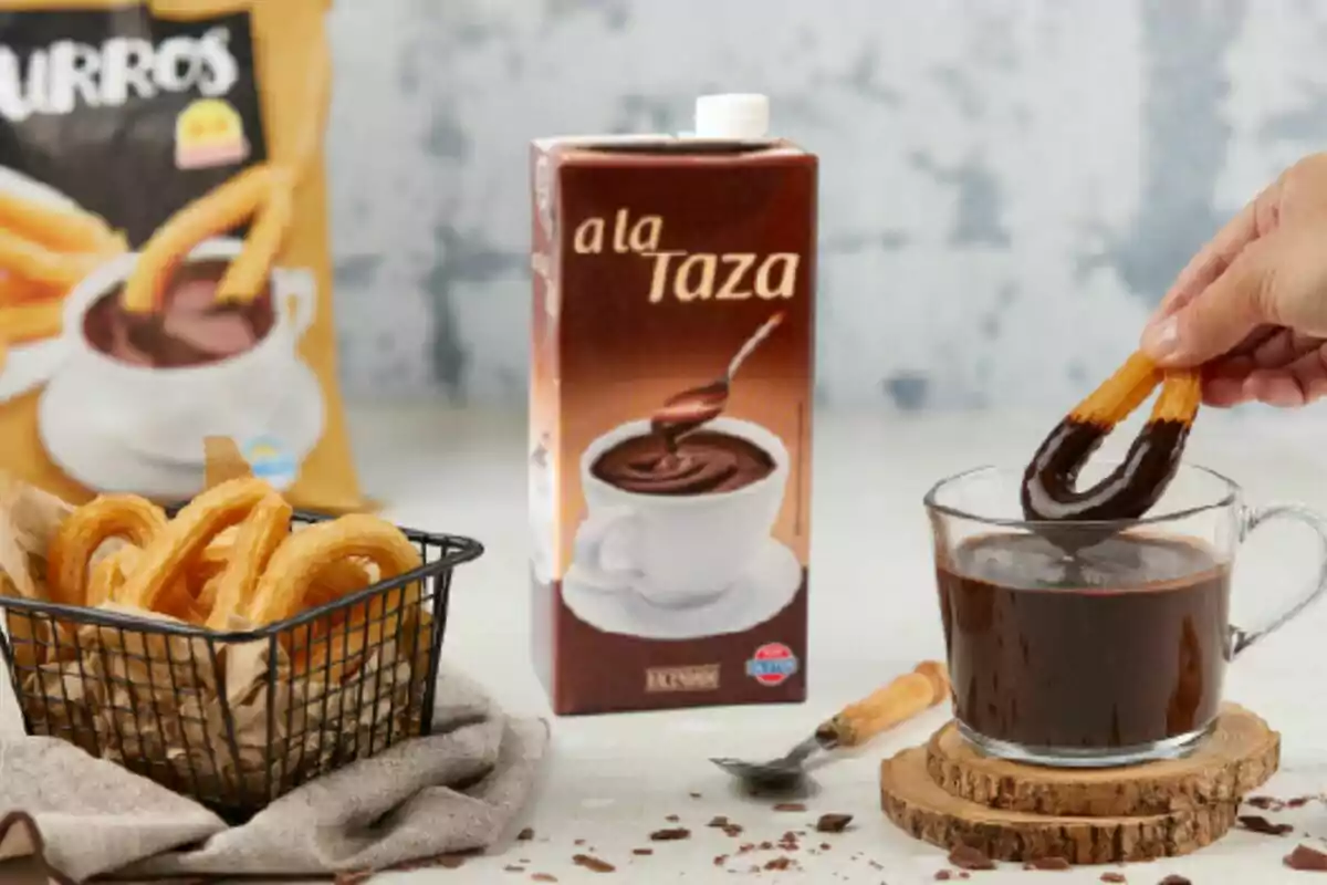 Una mano sumerge un churro en una taza de chocolate caliente junto a una caja de chocolate a la taza y una canasta con churros sobre una mesa Una mano sumerge un churro en una taza de chocolate caliente junto a una caja de chocolate a la taza y una canasta con churros sobre una mesa