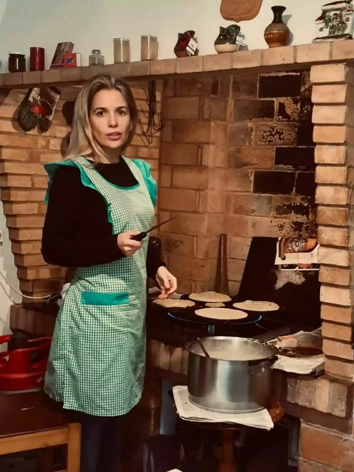 Christine Cambeiro con delantal verde cocina tortillas en una estufa de ladrillo, con una olla de metal en primer plano y varios objetos decorativos en la repisa superior. Post en Instagram el 7 de marzo de 2023