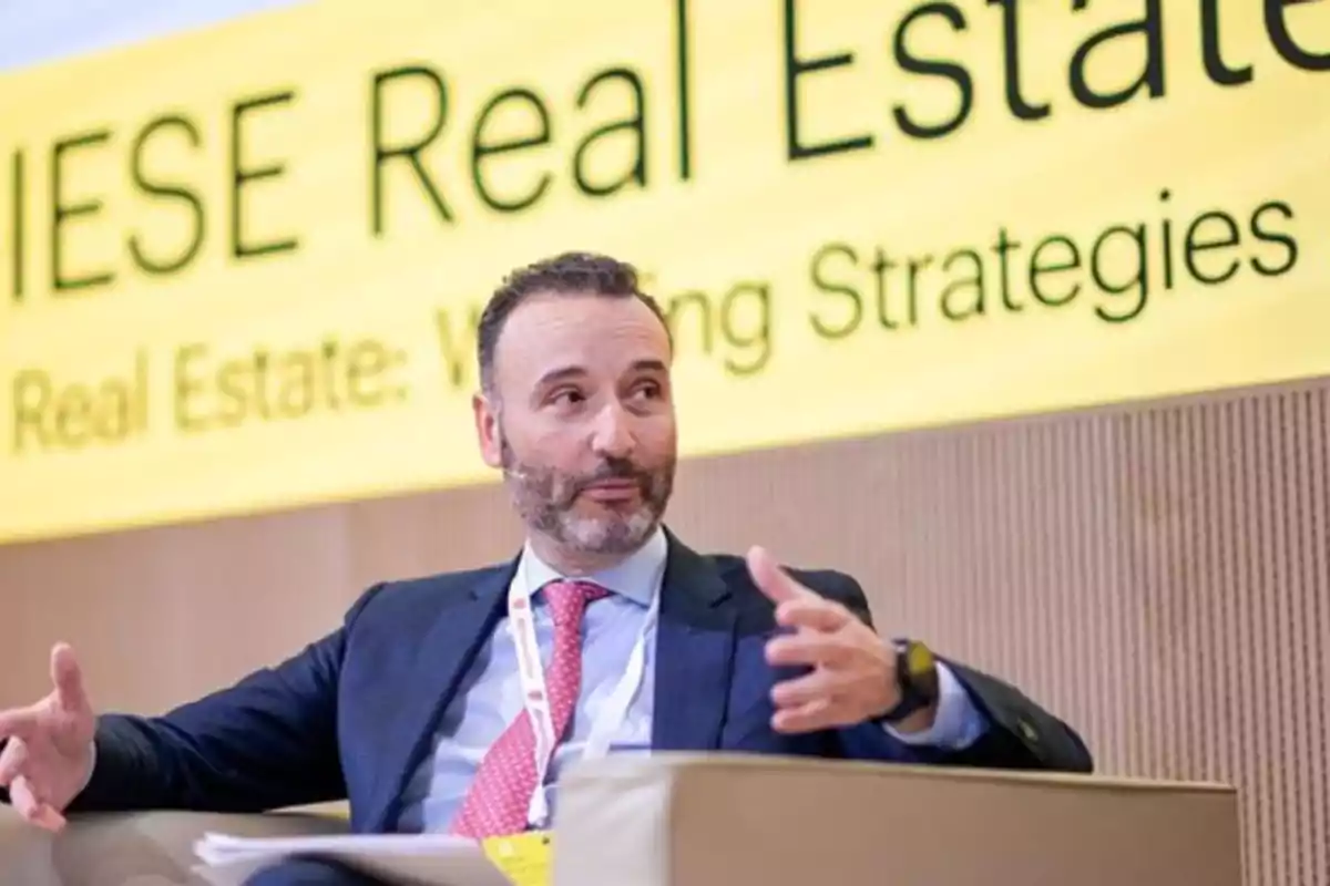 Hombre con barba y traje hablando en un panel de discusión sobre bienes raíces con un fondo amarillo que muestra texto relacionado al evento Hombre con barba y traje hablando en un panel de discusión sobre bienes raíces con un fondo amarillo que muestra texto relacionado al evento