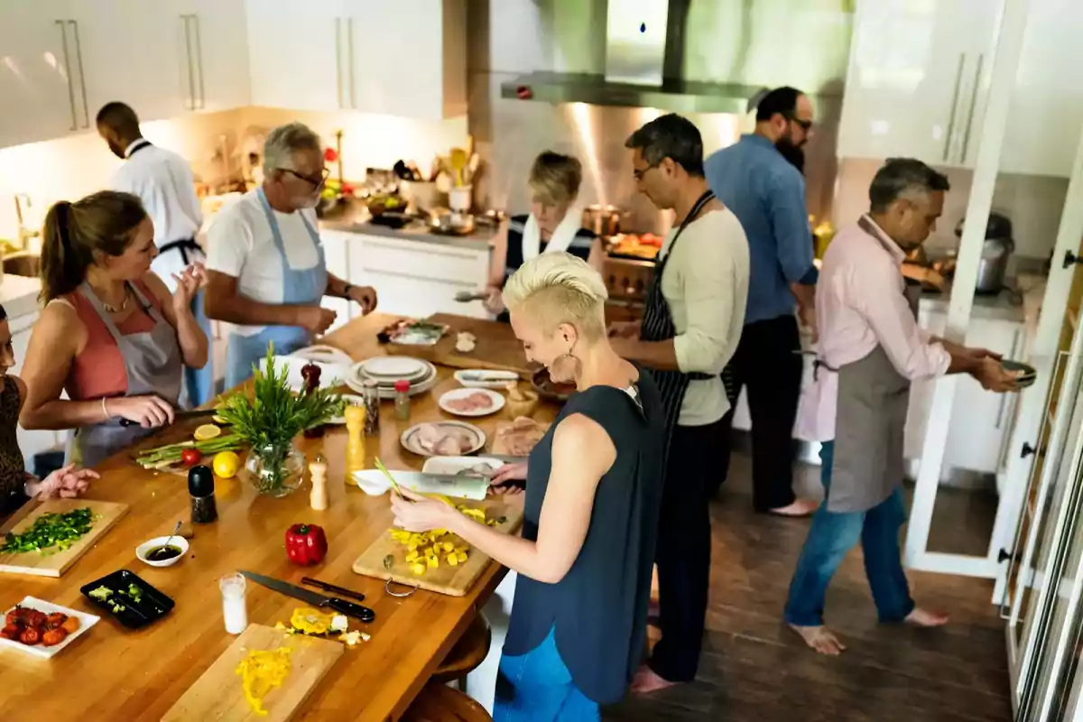 Un grupo de personas cocinando juntas en una cocina moderna, cada una preparando diferentes ingredientes en una mesa de madera.