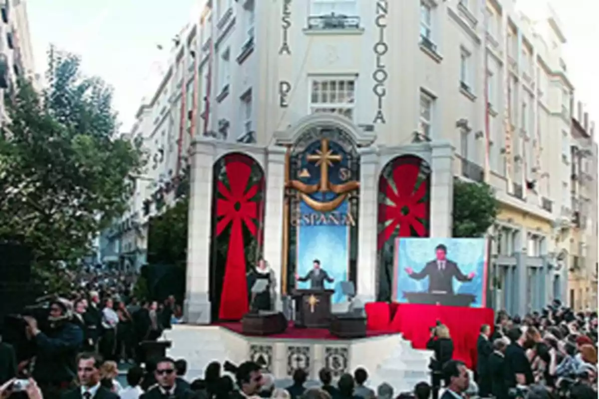 Una multitud se reúne frente a un edificio decorado con símbolos y banderas rojas, donde un orador se encuentra en un escenario con un podio y una pantalla grande mostrando su imagen.