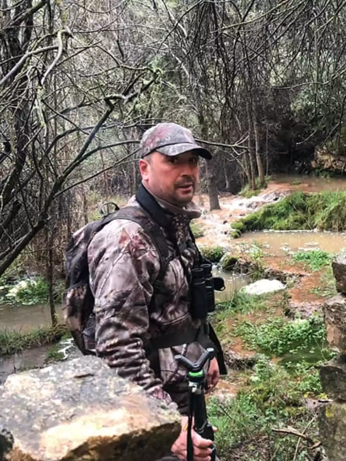 Hombre con ropa de camuflaje y gorra en un entorno boscoso junto a un arroyo.