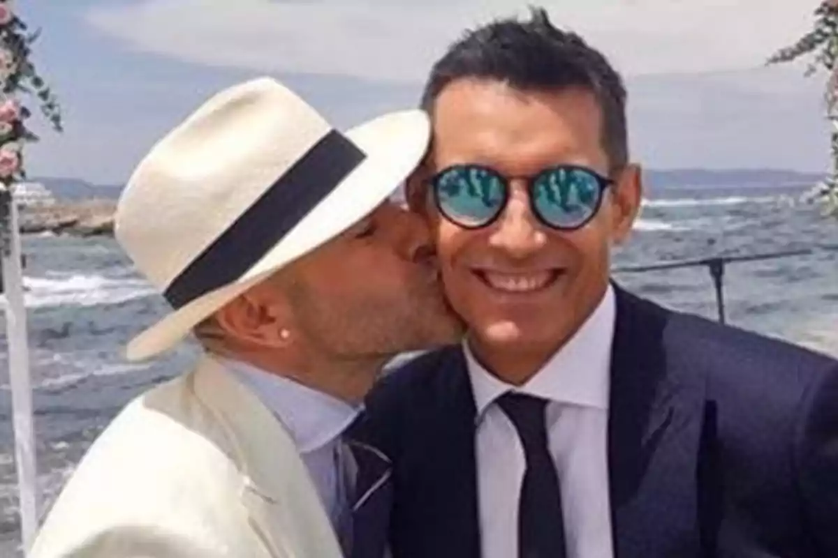 Dos hombres elegantemente vestidos sonríen mientras uno besa en la mejilla al otro junto al mar en un ambiente festivo Dos hombres elegantemente vestidos sonríen mientras uno besa en la mejilla al otro junto al mar en un ambiente festivo