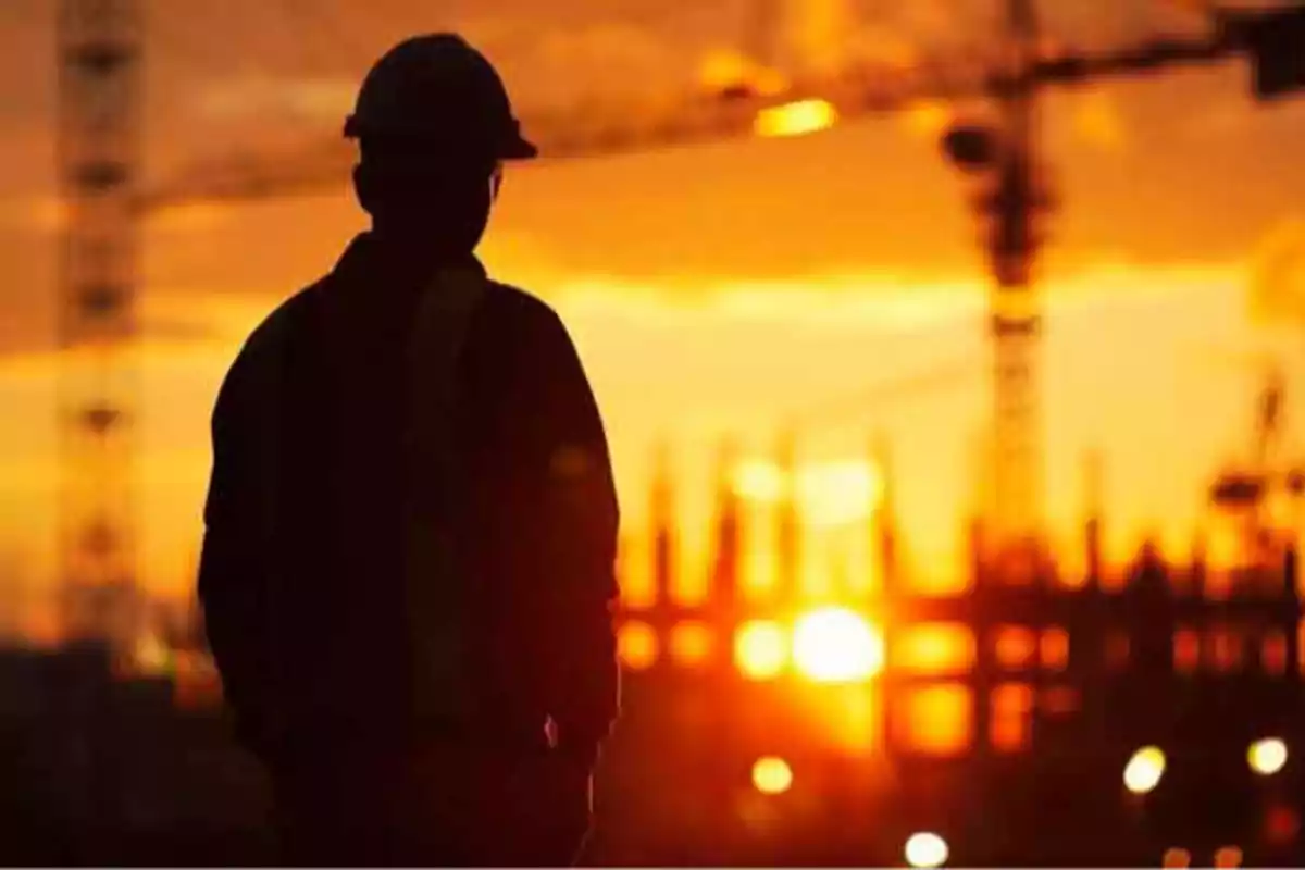 Trabajador de la construcción con casco observando un atardecer en un sitio de obra con grúas al fondo.