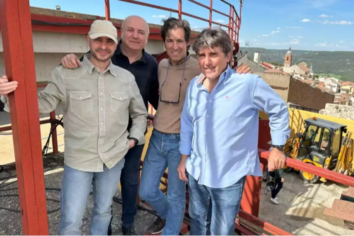 Cuatro hombres posan sonrientes en una estructura metálica al aire libre con un pueblo y una iglesia al fondo en un día soleado