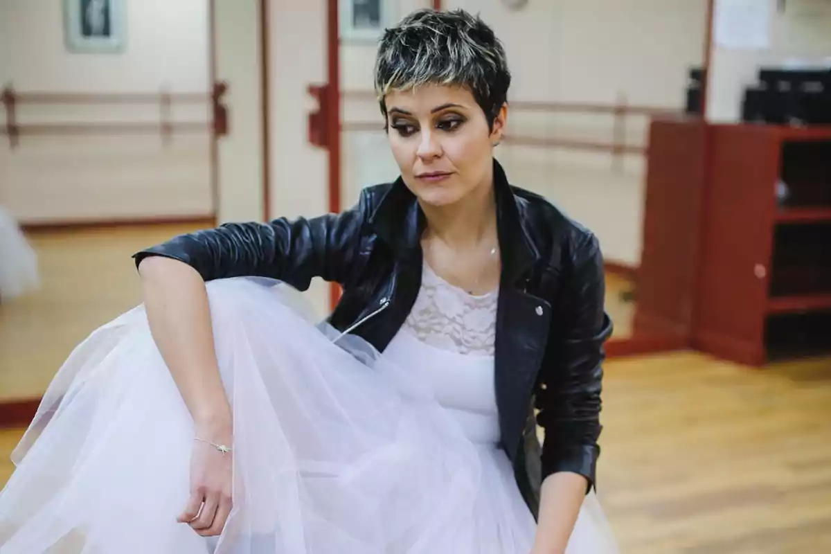 Mujer con vestido blanco de tul y chaqueta de cuero negra sentada en un estudio de danza con expresión pensativa Mujer con vestido blanco de tul y chaqueta de cuero negra sentada en un estudio de danza con expresión pensativa