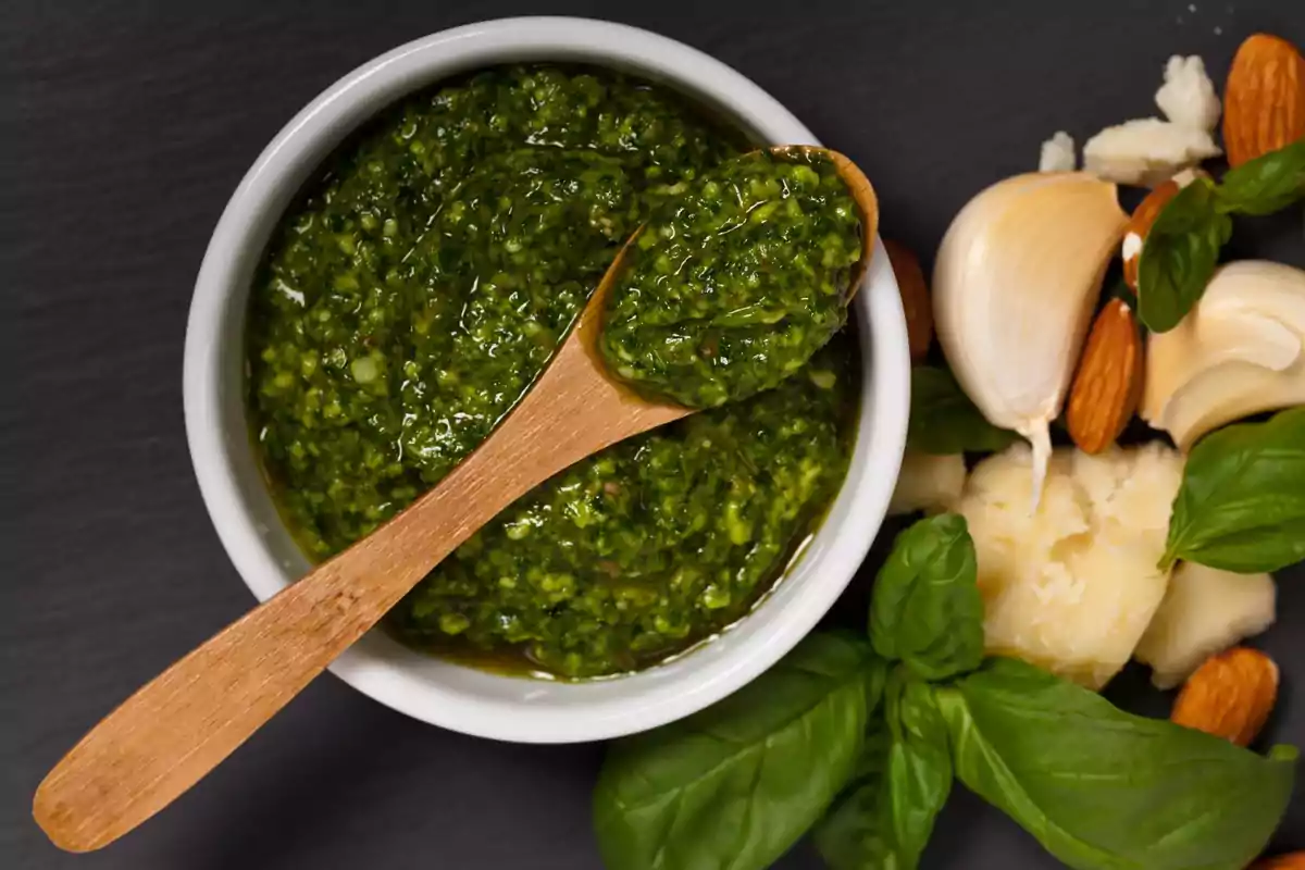 Un tazón de pesto con una cuchara de madera rodeado de ingredientes frescos como albahaca, ajo, almendras y queso.