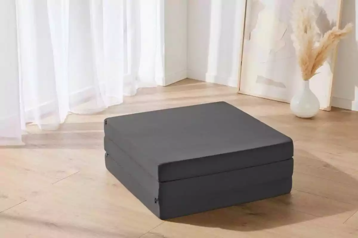 Un cojín cuadrado gris oscuro en el suelo de una habitación con piso de madera clara y cortinas blancas. Un cojín cuadrado gris oscuro en el suelo de una habitación con piso de madera clara y cortinas blancas.