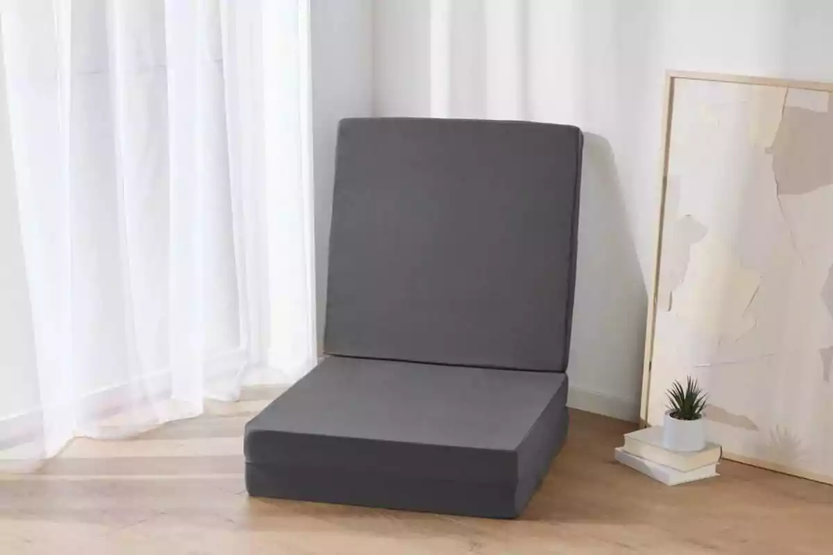 Una silla plegable gris en una habitación con suelo de madera, junto a una ventana con cortinas blancas y un cuadro apoyado en la pared. Una silla plegable gris en una habitación con suelo de madera, junto a una ventana con cortinas blancas y un cuadro apoyado en la pared.