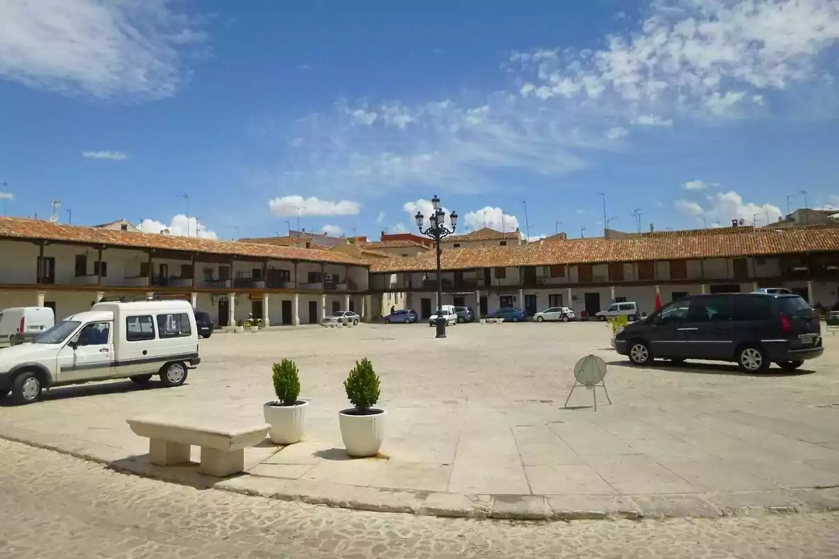 Plaza con coches estacionados rodeada de edificios antiguos y cielo azul con nubes.