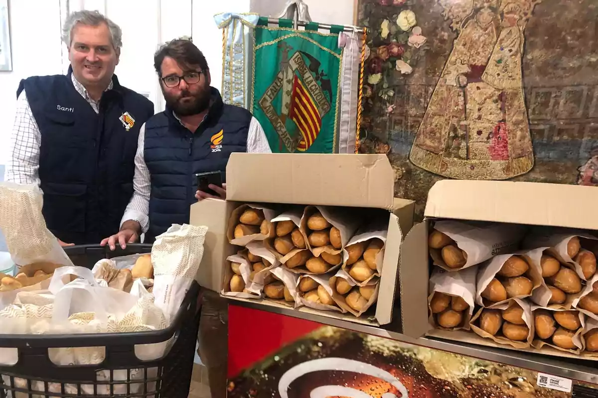 Dos personas posan junto a cajas llenas de panes en un entorno decorado con un estandarte y una imagen religiosa.