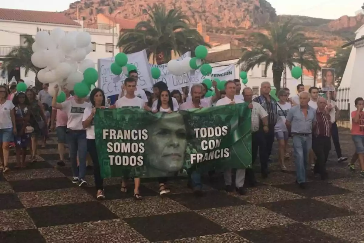 Un grupo de personas participa en una manifestación sosteniendo pancartas y globos verdes y blancos en un entorno al aire libre con palmeras y montañas al fondo.