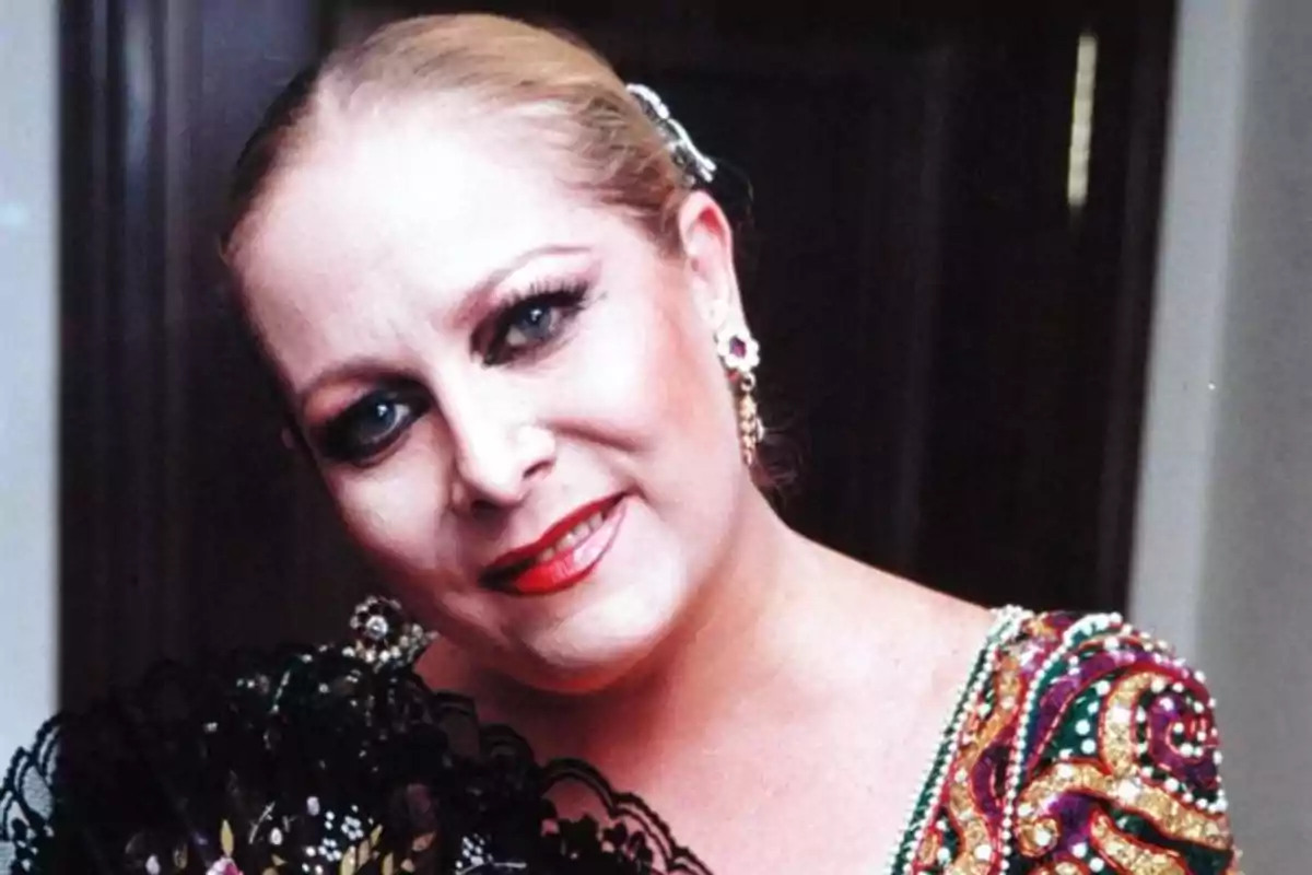 Mujer rubia con maquillaje llamativo, vestido colorido y abanico negro decorado, sonriendo frente a una puerta oscura Mujer rubia con maquillaje llamativo, vestido colorido y abanico negro decorado, sonriendo frente a una puerta oscura