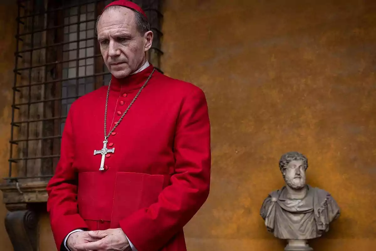 Un hombre vestido con sotana roja y birrete, con un crucifijo colgando, está de pie frente a una pared de color ocre junto a un busto de mármol. Un hombre vestido con sotana roja y birrete, con un crucifijo colgando, está de pie frente a una pared de color ocre junto a un busto de mármol.