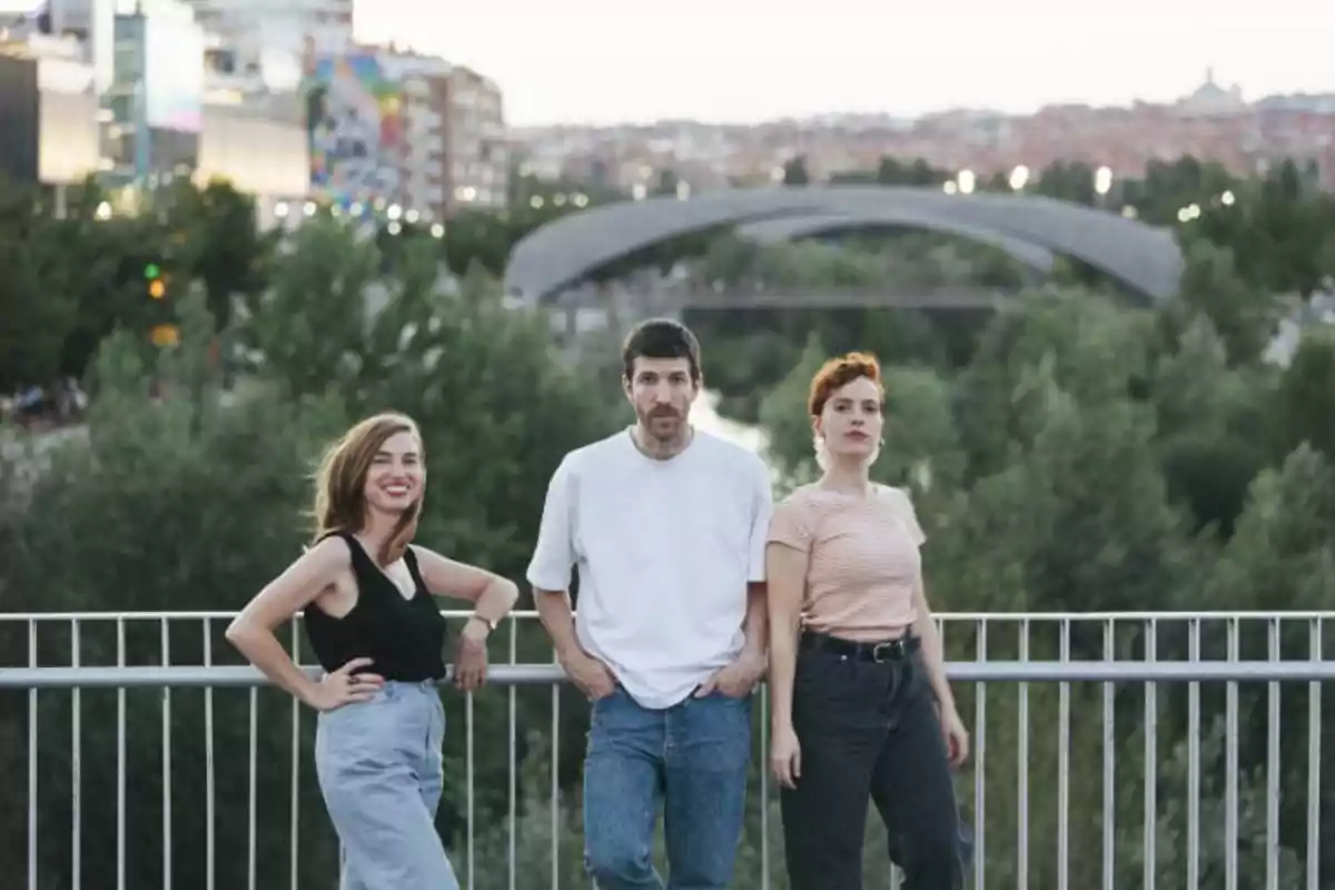 Tres personas posan sobre un puente con un paisaje urbano y un puente de fondo.