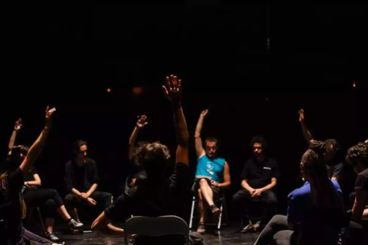 Un grupo de personas sentadas en círculo levantando las manos en un ambiente oscuro.