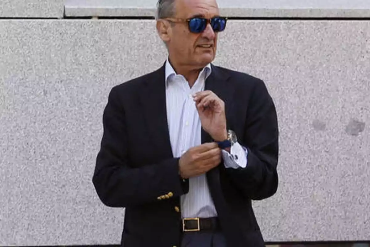 Hombre con gafas de sol y traje oscuro ajustándose el reloj frente a una pared de piedra.