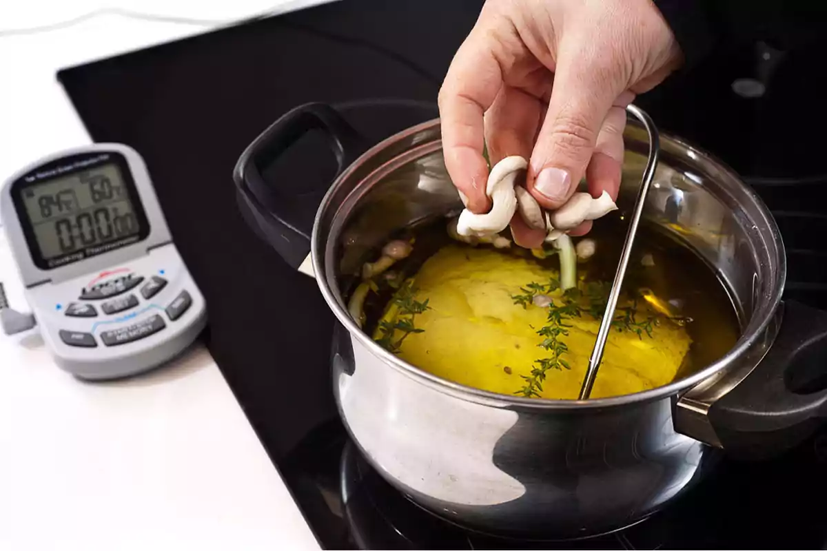 Una mano agregando champiñones a una olla con líquido y hierbas, con un termómetro y un temporizador de cocina al lado.