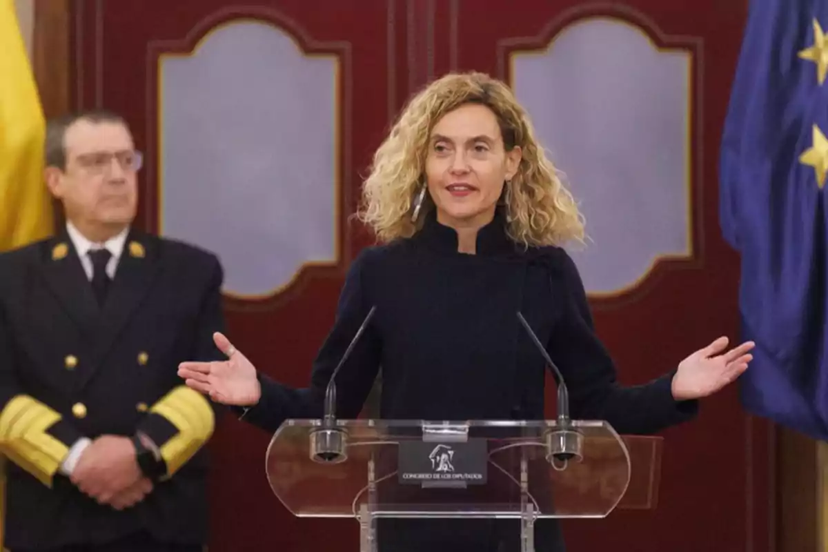 Una mujer de cabello rizado y rubio habla en un podio con un hombre uniformado al fondo. Una mujer de cabello rizado y rubio habla en un podio con un hombre uniformado al fondo.