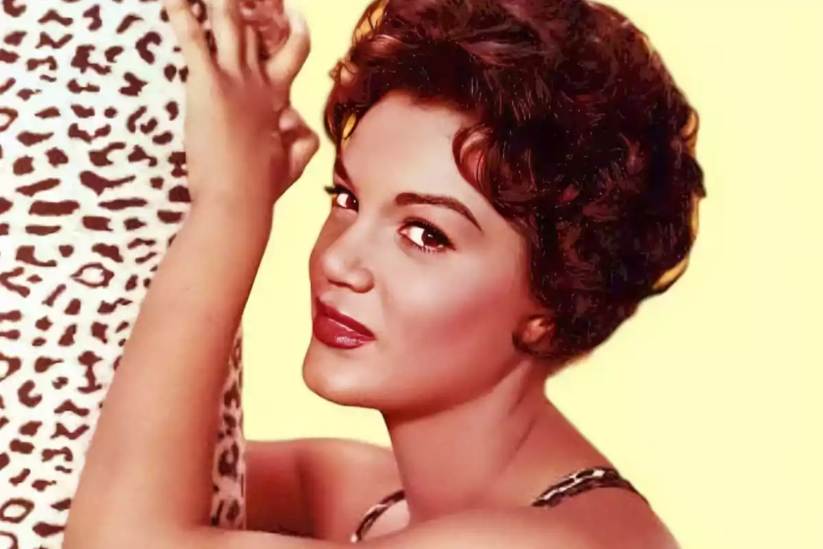 Mujer con cabello corto y rizado mirando a la cámara mientras sostiene una tela con estampado de leopardo sobre un fondo amarillo.