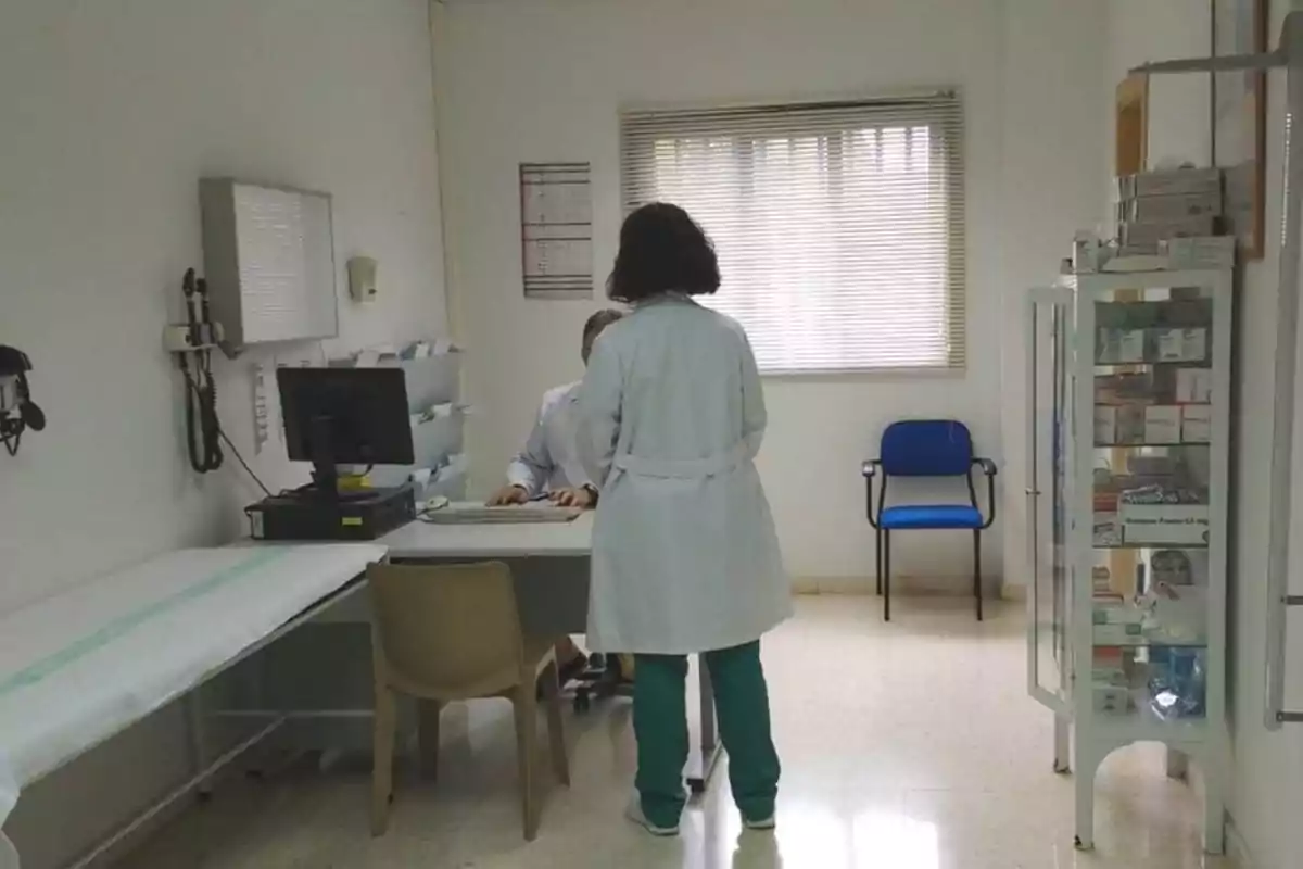 Consultorio médico con dos personas, una sentada frente a una computadora y otra de pie con bata blanca.