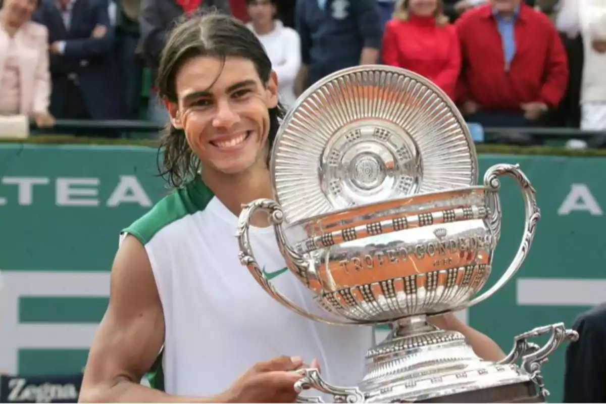 Un tenista sonriente sostiene un gran trofeo plateado en una cancha de tenis con espectadores al fondo.