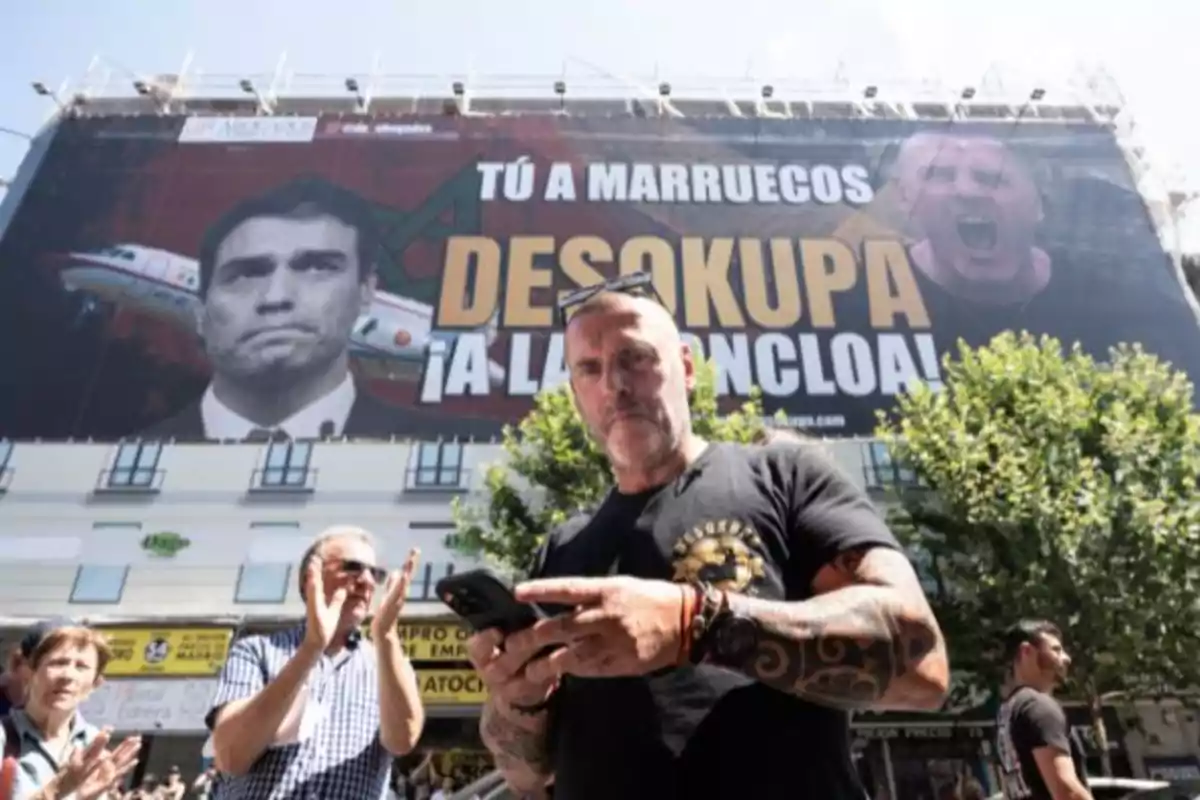 Un hombre con tatuajes sostiene un teléfono frente a un gran cartel publicitario en un edificio, mientras otras personas aplauden a su alrededor.